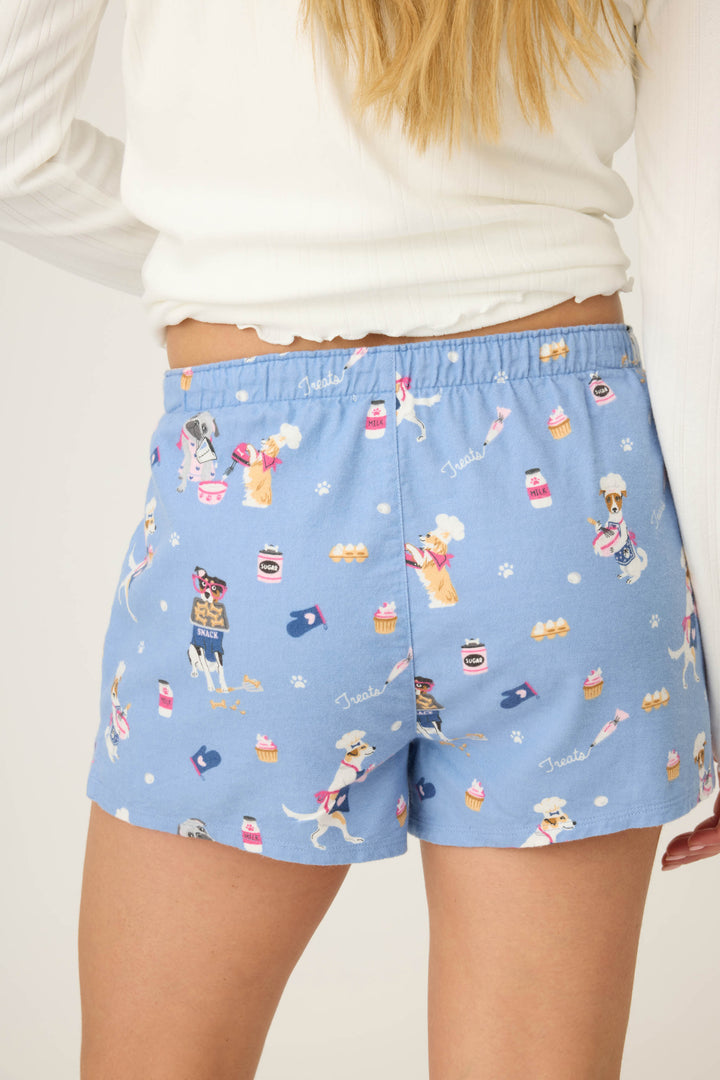 A person is seen from the back wearing P.J. Salvage DOGS SHORT FLANNELS, blue pajama shorts with a playful dog and bakery-themed print, paired with a white long-sleeve top.