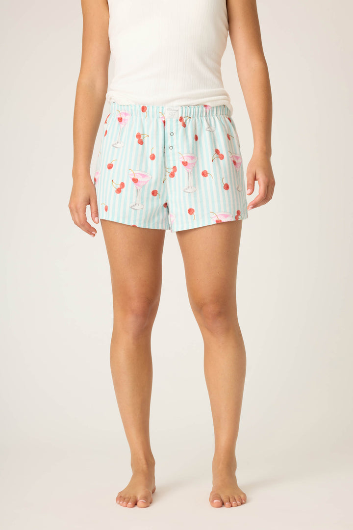 A person stands barefoot in P.J. Salvage SHORT FLANNELS—light blue and white striped shorts with a pink cocktail and cherry print—paired with a white sleeveless top. Arms relaxed, they pose against a plain light background.