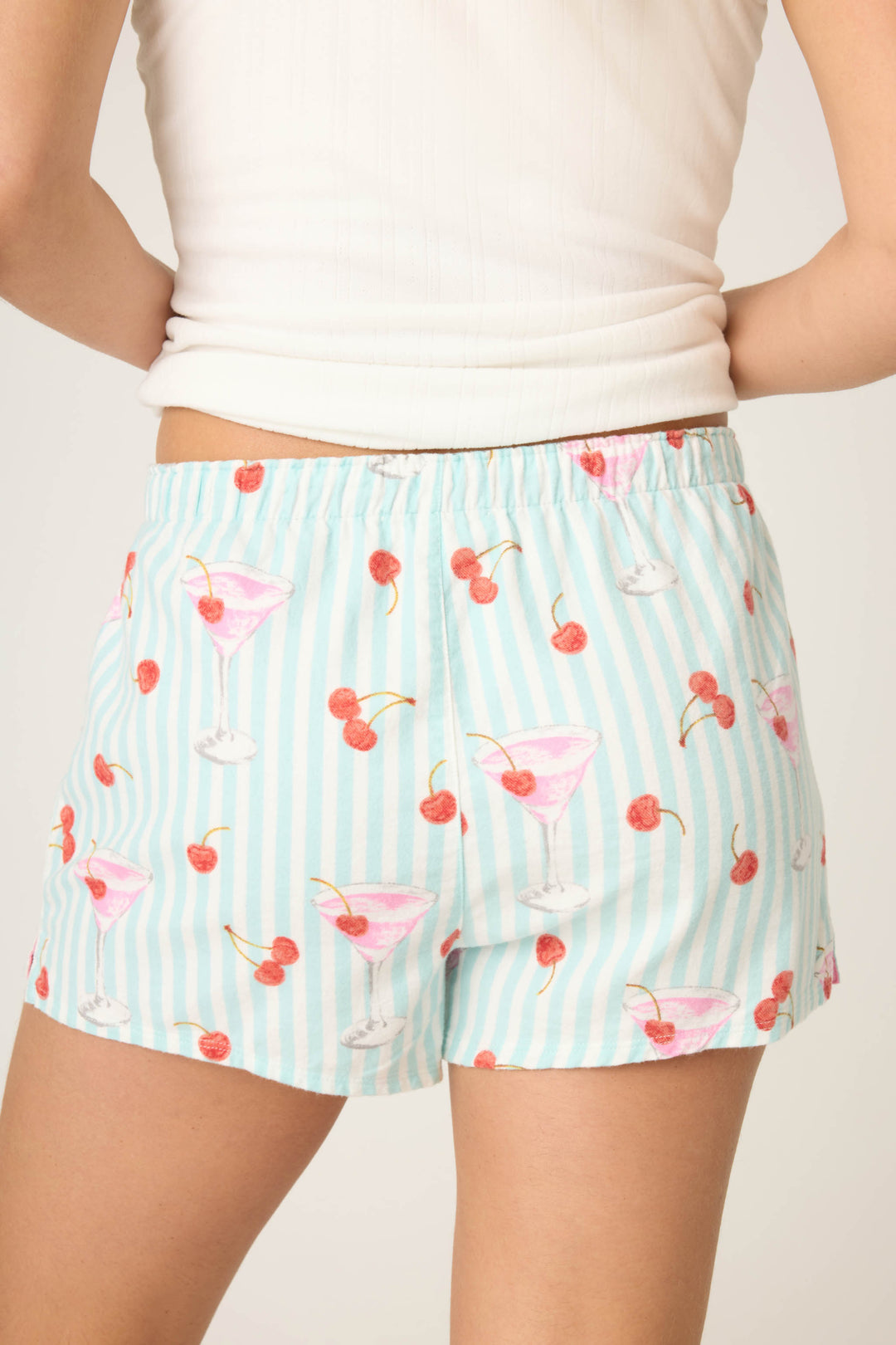 A back view from the waist down shows a person in P.J. Salvage SHORT FLANNELS—light blue and white striped shorts decorated with pink cocktail glasses and red cherries—paired with a white sleeveless top.