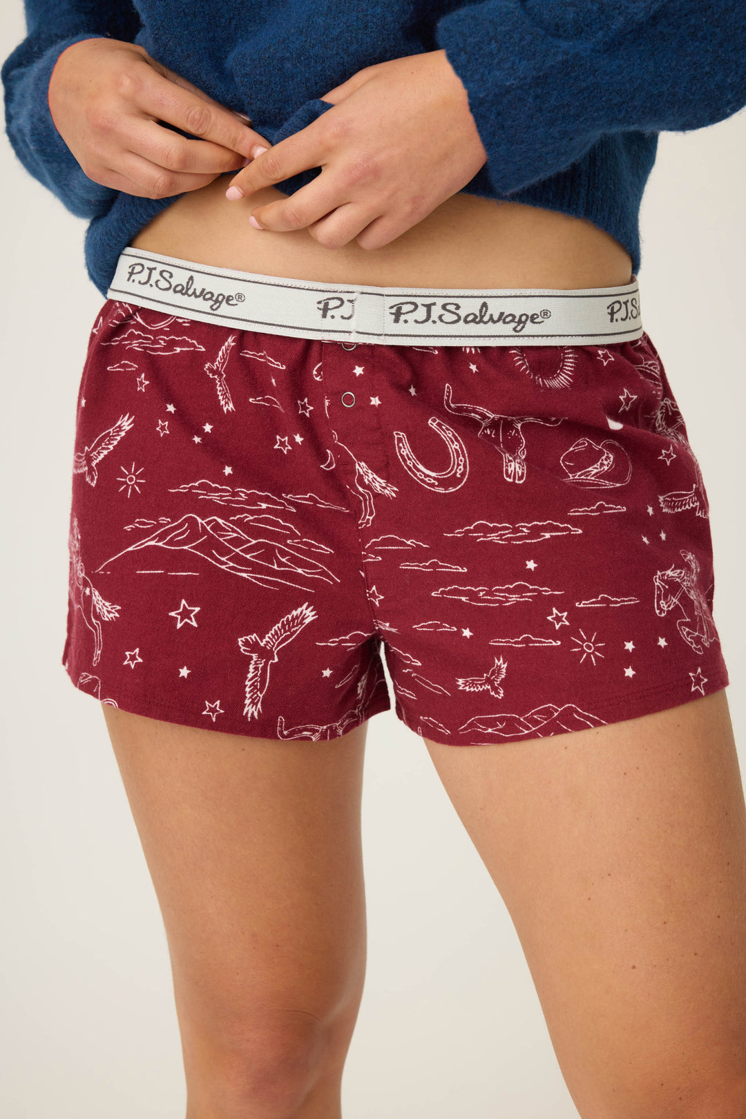 A person wearing maroon P.J. Salvage SHORT FLANNELS with white western-themed line drawings and a blue sweater. The photo is cropped to show only the lower torso and upper legs.