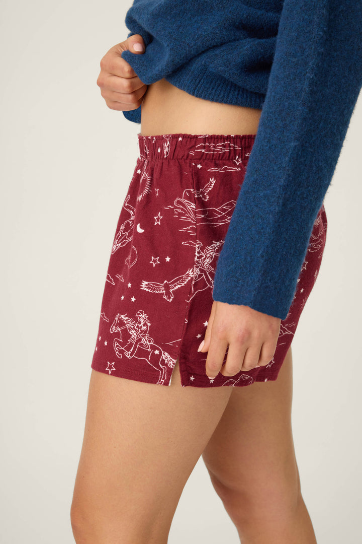 A person stands in a neutral pose wearing P.J. Salvage SHORT FLANNELS—red shorts featuring a white celestial and horse print—with a blue long-sleeve sweater, against a plain background.