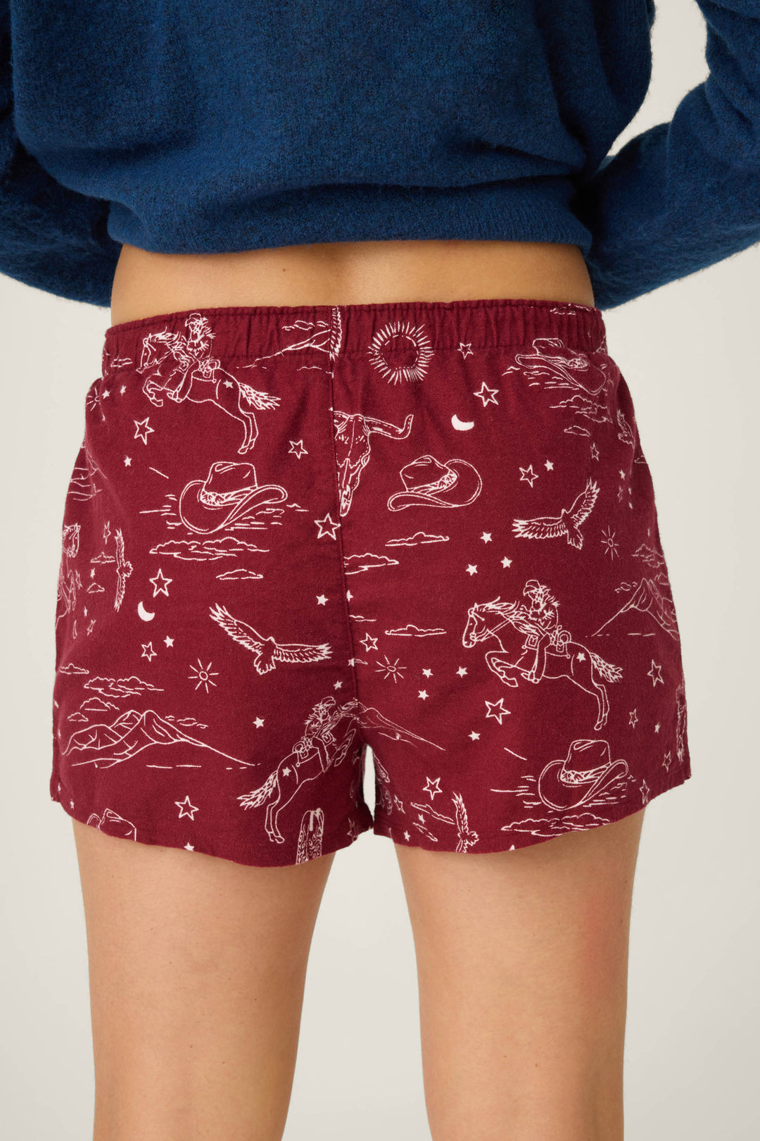 A person wears P.J. Salvage SHORT FLANNELS—maroon shorts featuring white western-themed illustrations (horses, cowboys, stars, and animals). The photo is taken from behind and also shows part of a dark blue top.