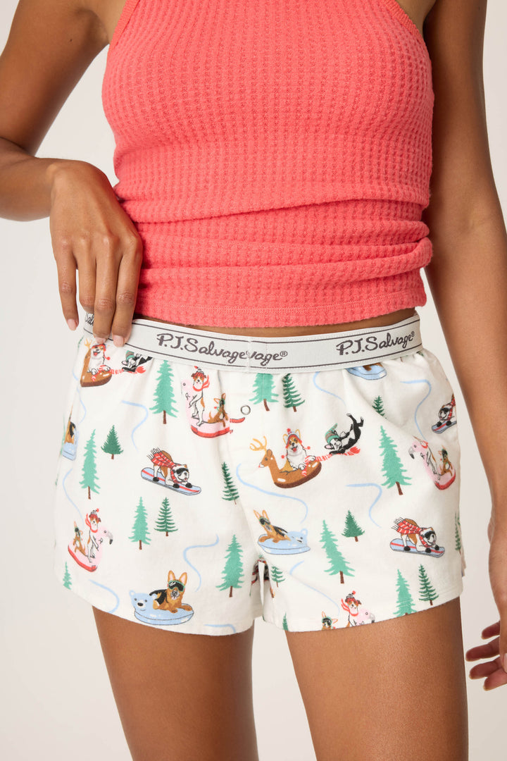 A person wears a pink textured tank top and P.J. Salvage Sleigh All Day Winter Print Flannel Shorts with a playful dog sledding print, standing against a neutral background. The shorts’ waistband displays the P.J. Salvage brand name.