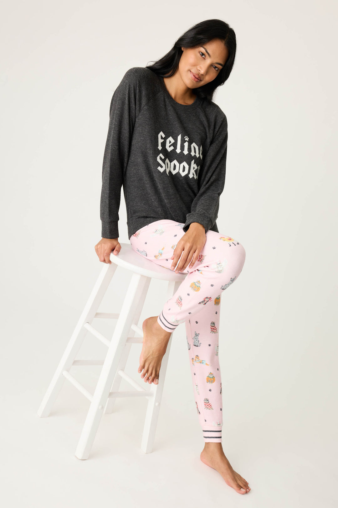 A person sits on a white stool, wearing the P.J. Salvage FEELINE SPOOKY LONG SLEEVE FELINE SPOOKY and light pink patterned pajama pants, set against a plain light background.