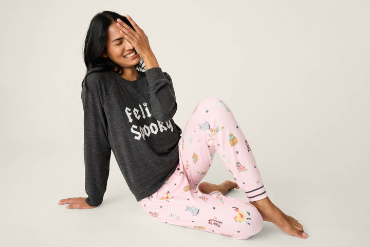 A woman sits on the floor smiling with one hand partly covering her face, wearing a dark gray P.J. Salvage FEELINE SPOOKY LONG SLEEVE FELINE SPOOKY shirt and light pink pajama pants with colorful cartoon prints.