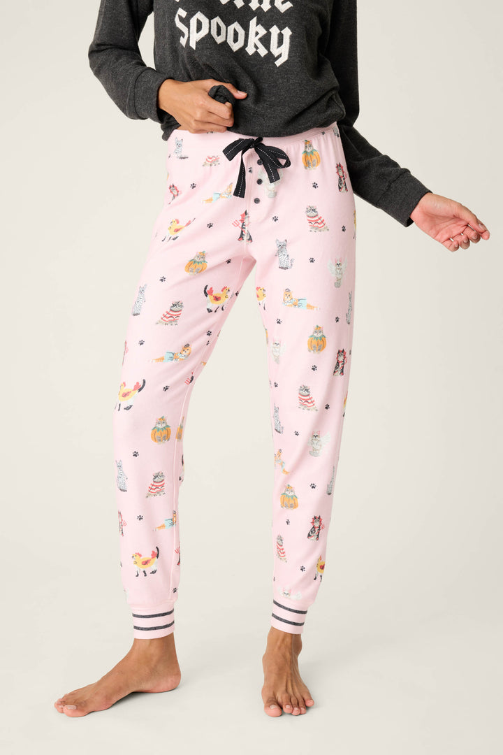 A person wears P.J. Salvage CATS JAMMIE PANT FELINE SPOOKY—pink pajama pants with a colorful animal print and black drawstring—paired with a black long-sleeve Spooky shirt; barefoot.