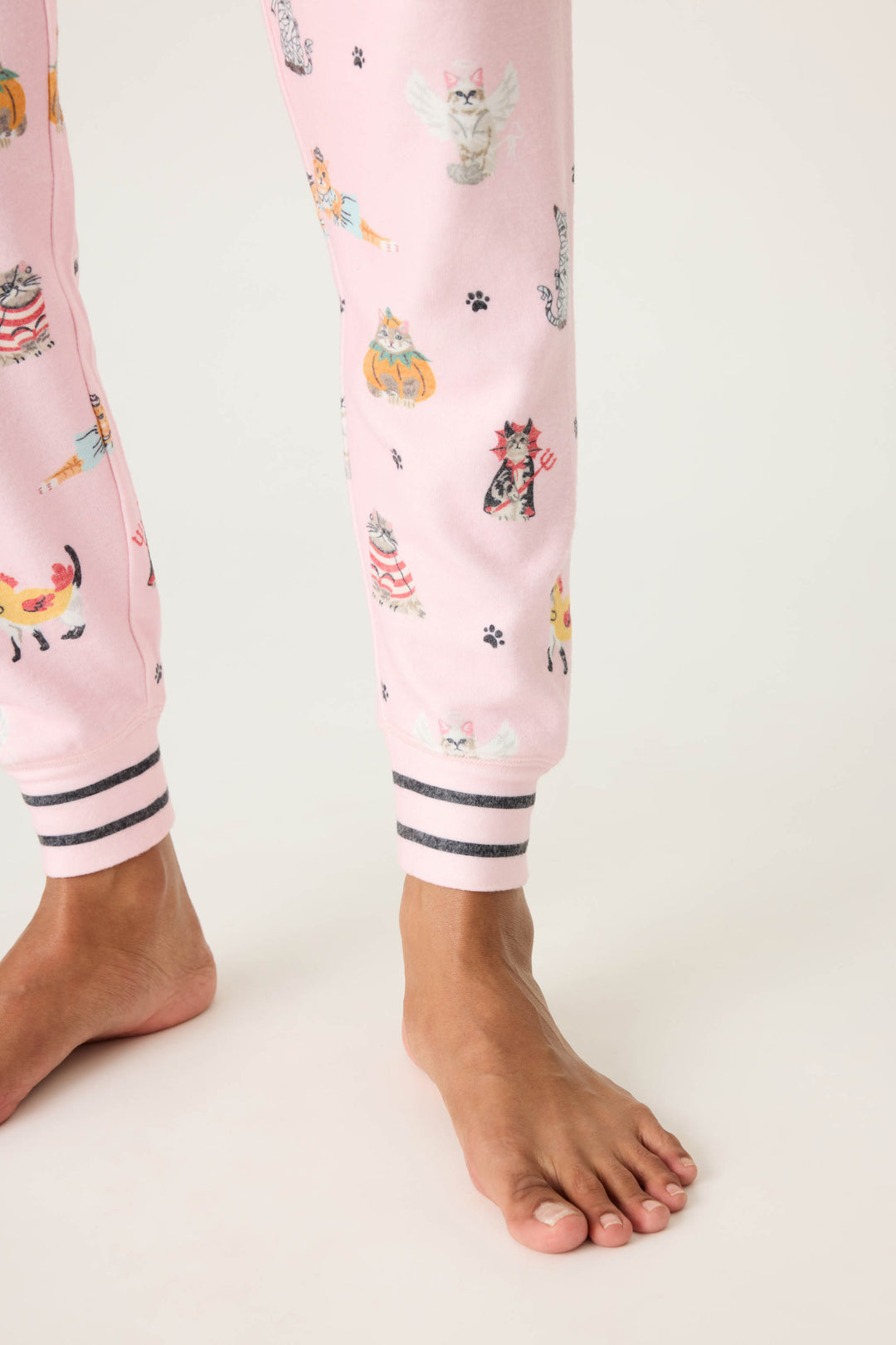 A person stands barefoot on a light surface, wearing P.J. Salvage CATS JAMMIE PANT FELINE SPOOKY pajama pants with cartoon cat prints and black-and-white striped ankle cuffs.