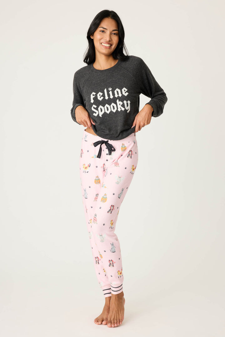 A person stands barefoot, smiling and holding the hem of a dark grey P.J. Salvage sweatshirt with Feline Spooky text, paired with CATS JAMMIE PANT FELINE SPOOKY light pink pajama pants featuring cartoon prints.
