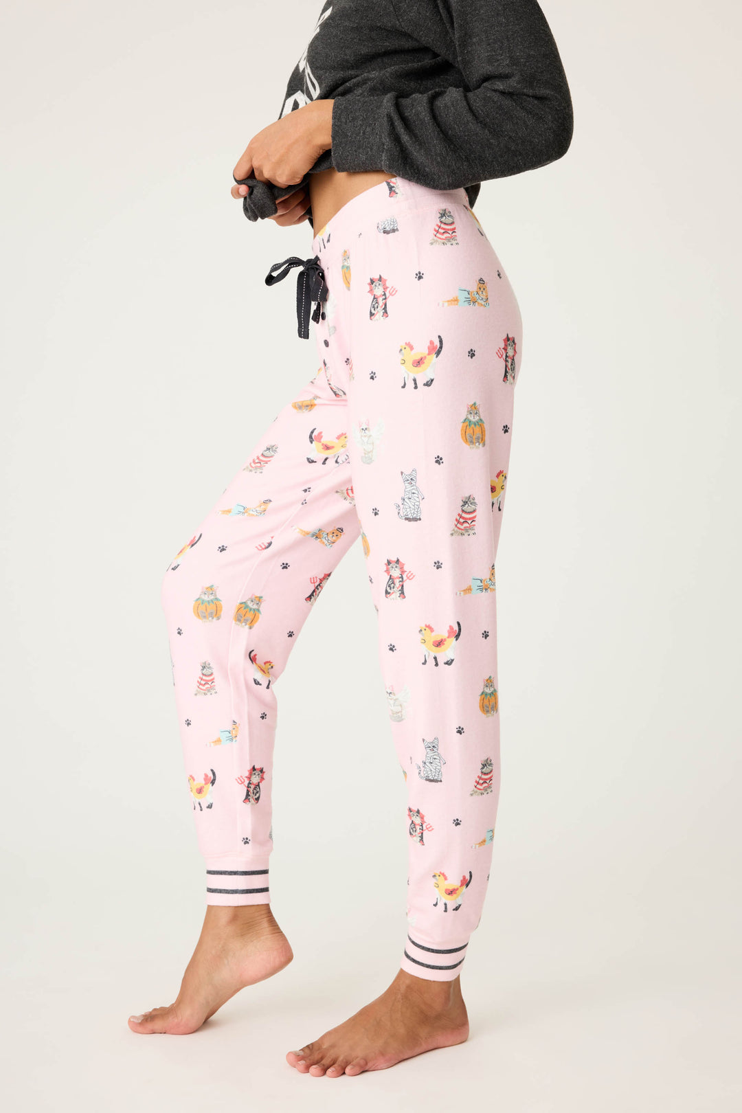 A person stands barefoot on one foot wearing P.J. Salvage CATS JAMMIE PANT FELINE SPOOKY pajama pants with colorful animal and cupcake print, black drawstrings, and striped cuffs. Their dark sweatshirt is partially visible.