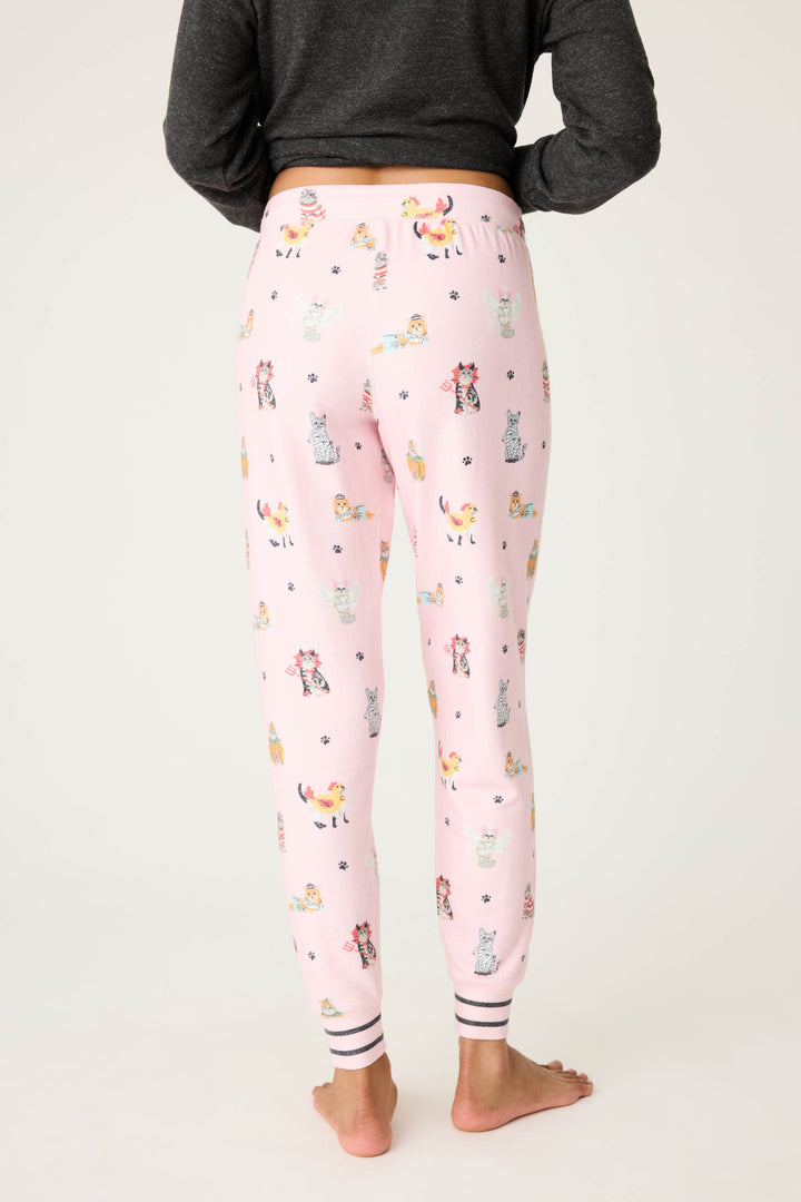 A person is seen from the back, barefoot, wearing P.J. Salvage CATS JAMMIE PANT FELINE SPOOKY—light pink pajama pants with a playful cat print and white-black striped ankle cuffs—paired with a dark long-sleeve top.