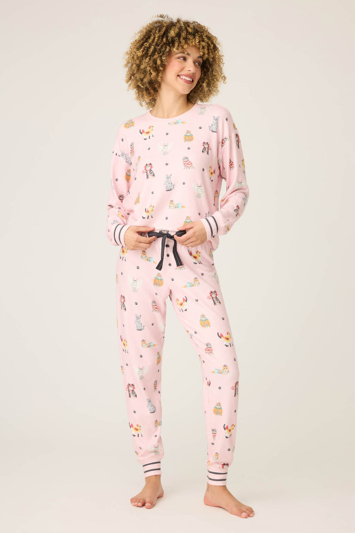 A person stands barefoot in the P.J. Salvage CATS PJ SET FELINE SPOOKY, a pink pajama set with colorful cartoon designs, hands in pockets and looking to the side with a slight smile against a plain light background.