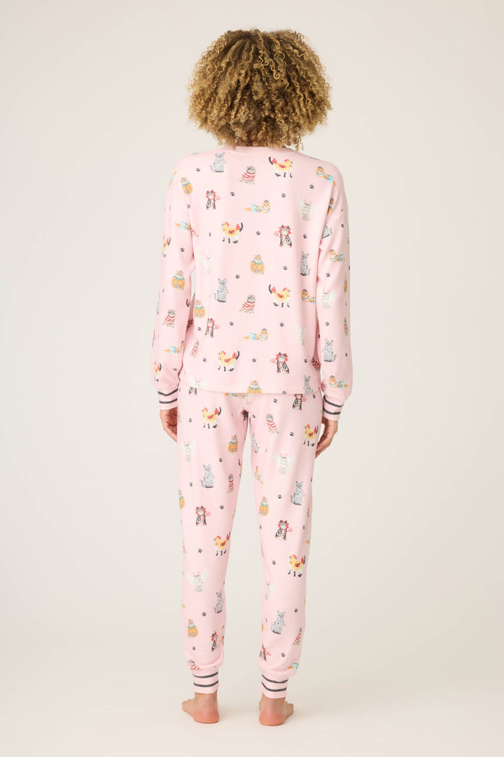 A person with curly hair stands barefoot, facing away from the camera, wearing the P.J. Salvage CATS PJ SET FELINE SPOOKY—light pink pajamas with a multicolored animal print—against a plain off-white background.