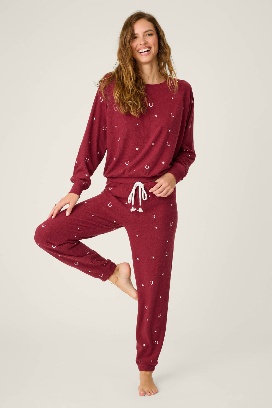 A woman models the P.J. Salvage Pant Set HORSE SHOE PJ SET HOLD YOUR HORSES—maroon pajamas with a white horseshoe pattern, drawstring waist, and long sleeves—standing on one leg and smiling against a plain light background.