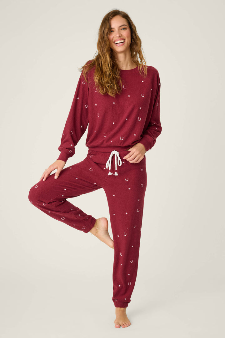 A woman models the P.J. Salvage Pant Set HORSE SHOE PJ SET HOLD YOUR HORSES—maroon pajamas with a white horseshoe pattern, drawstring waist, and long sleeves—standing on one leg and smiling against a plain light background.