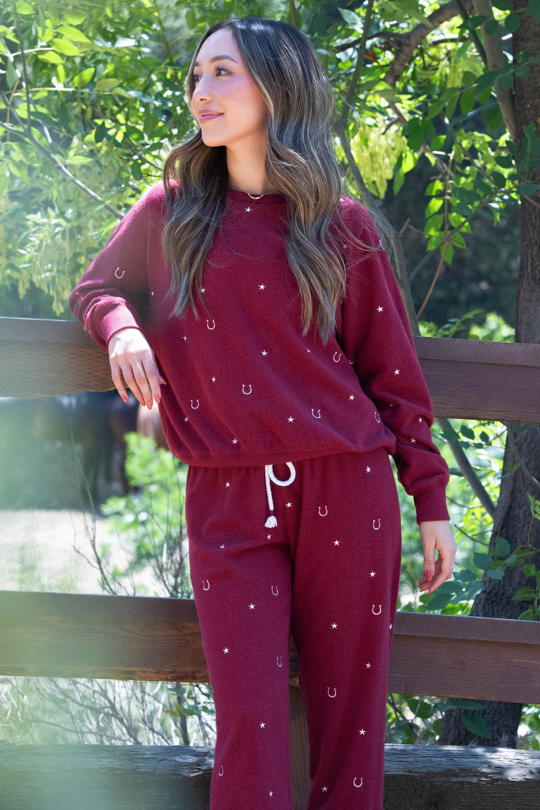 A woman stands outdoors by a wooden fence, wearing the P.J. Salvage Pant Set HORSE SHOE PJ SET HOLD YOUR HORSES in maroon with small white horseshoes and stars. She has long, wavy hair and gazes to the side amid green foliage.