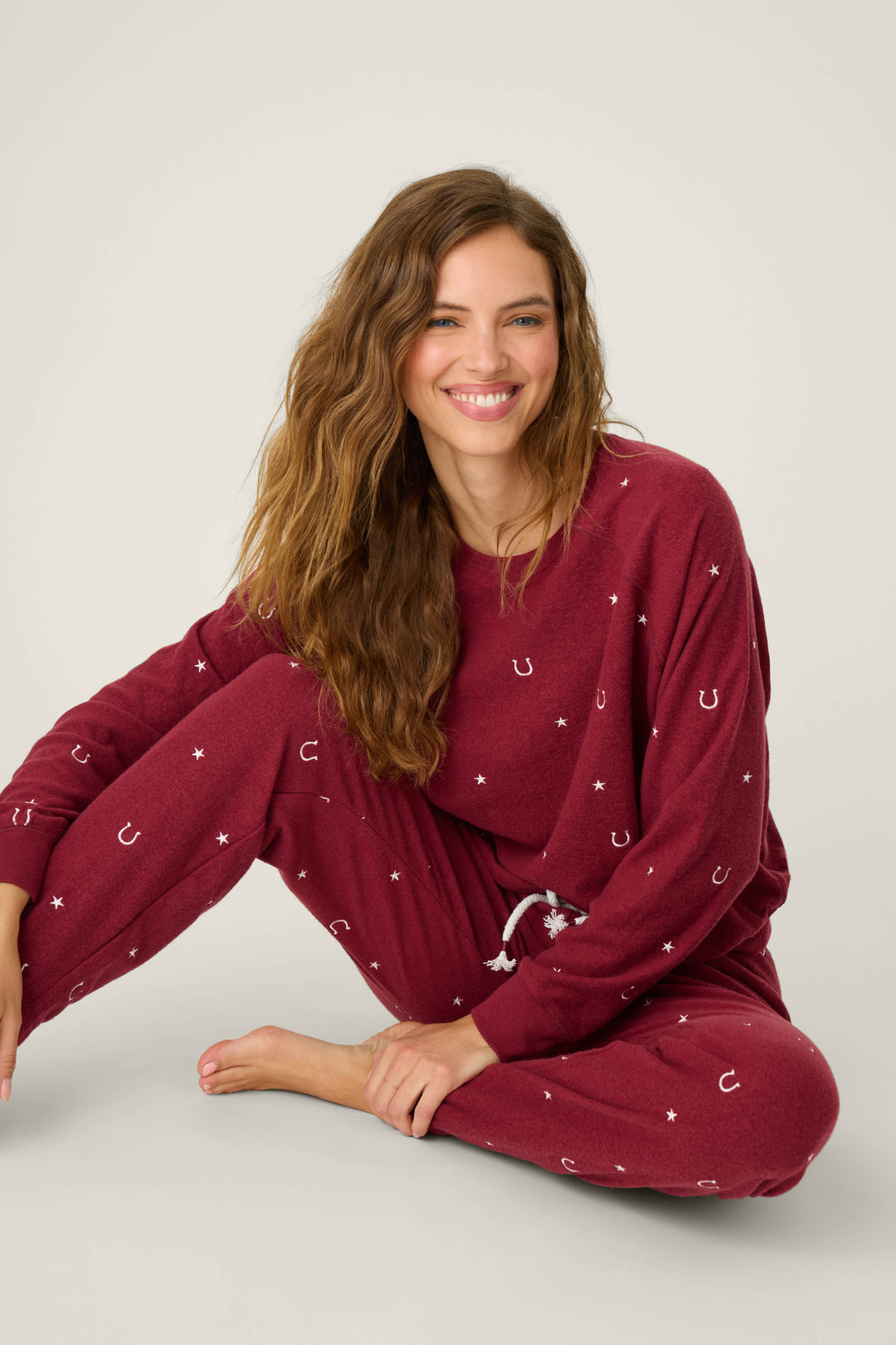 A woman with long wavy hair smiles on the floor, wearing the P.J. Salvage Pant Set HORSE SHOE PJ SET HOLD YOUR HORSES— a matching maroon pajama set with white horseshoe and star patterns, against a plain light background.