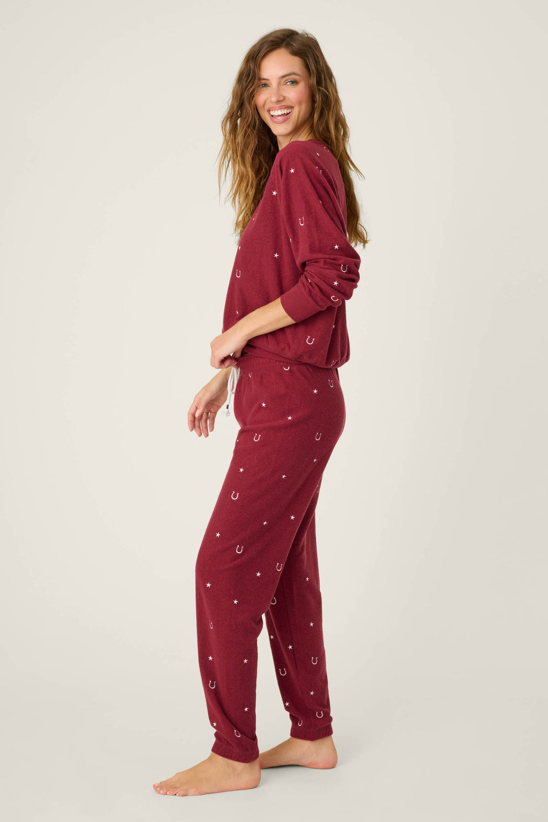 A woman, barefoot against a plain background, wears the P.J. Salvage Pant Set HORSE SHOE PJ SET HOLD YOUR HORSES in maroon with a small horseshoe print. She smiles at the camera, body turned slightly to the side.