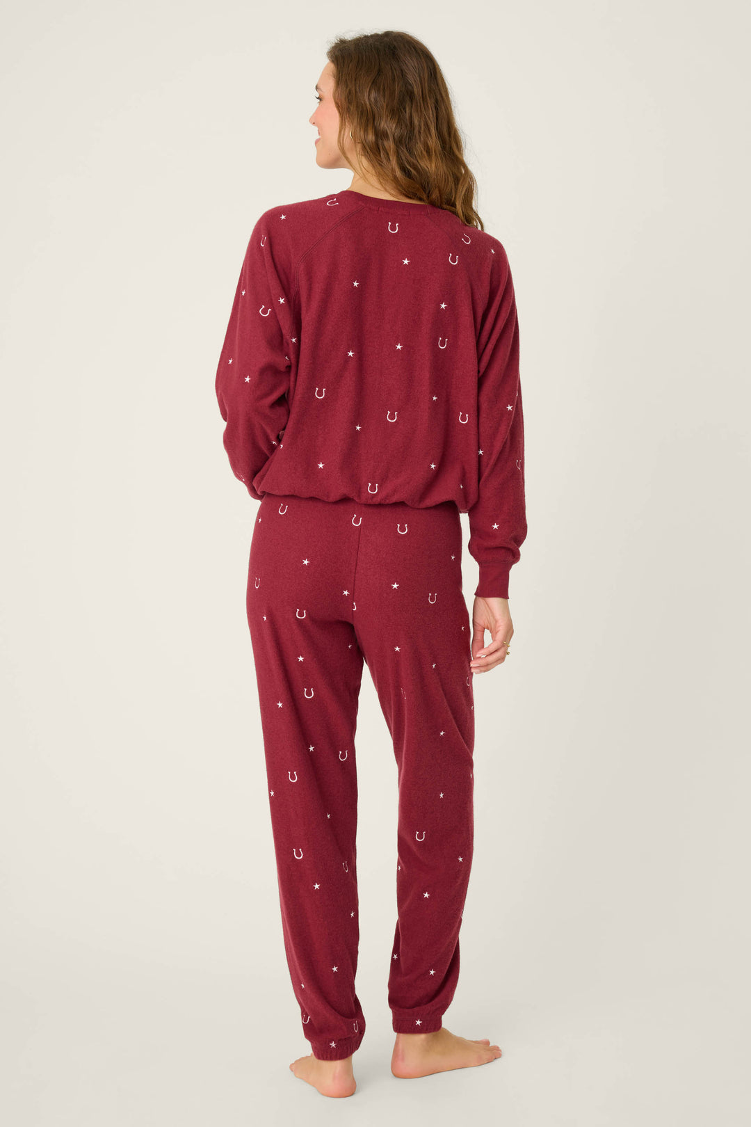A person stands barefoot facing away, wearing the P.J. Salvage Pant Set HORSE SHOE PJ SET HOLD YOUR HORSES, a matching maroon pajama set with small white horseshoe patterns, against a plain light-colored background.