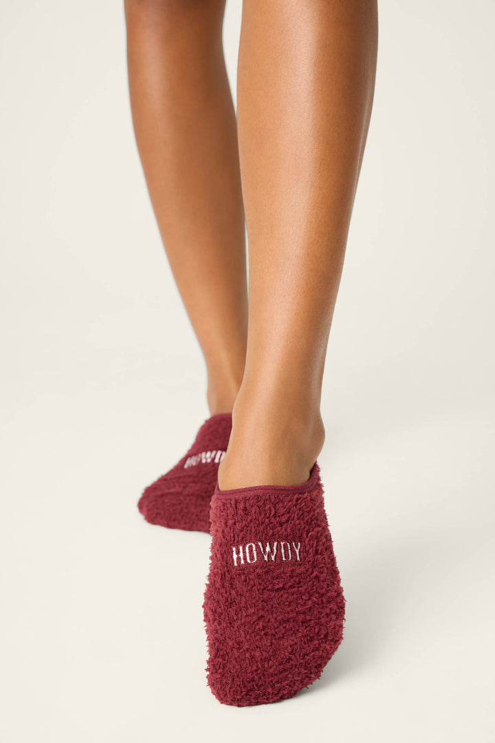 A person stands on a light surface wearing P.J. Salvage check color - HOWDY SOCKS FLANNELS, featuring fuzzy maroon fabric with HOWDY embroidered in white on the heels. Only the feet and lower legs are visible.