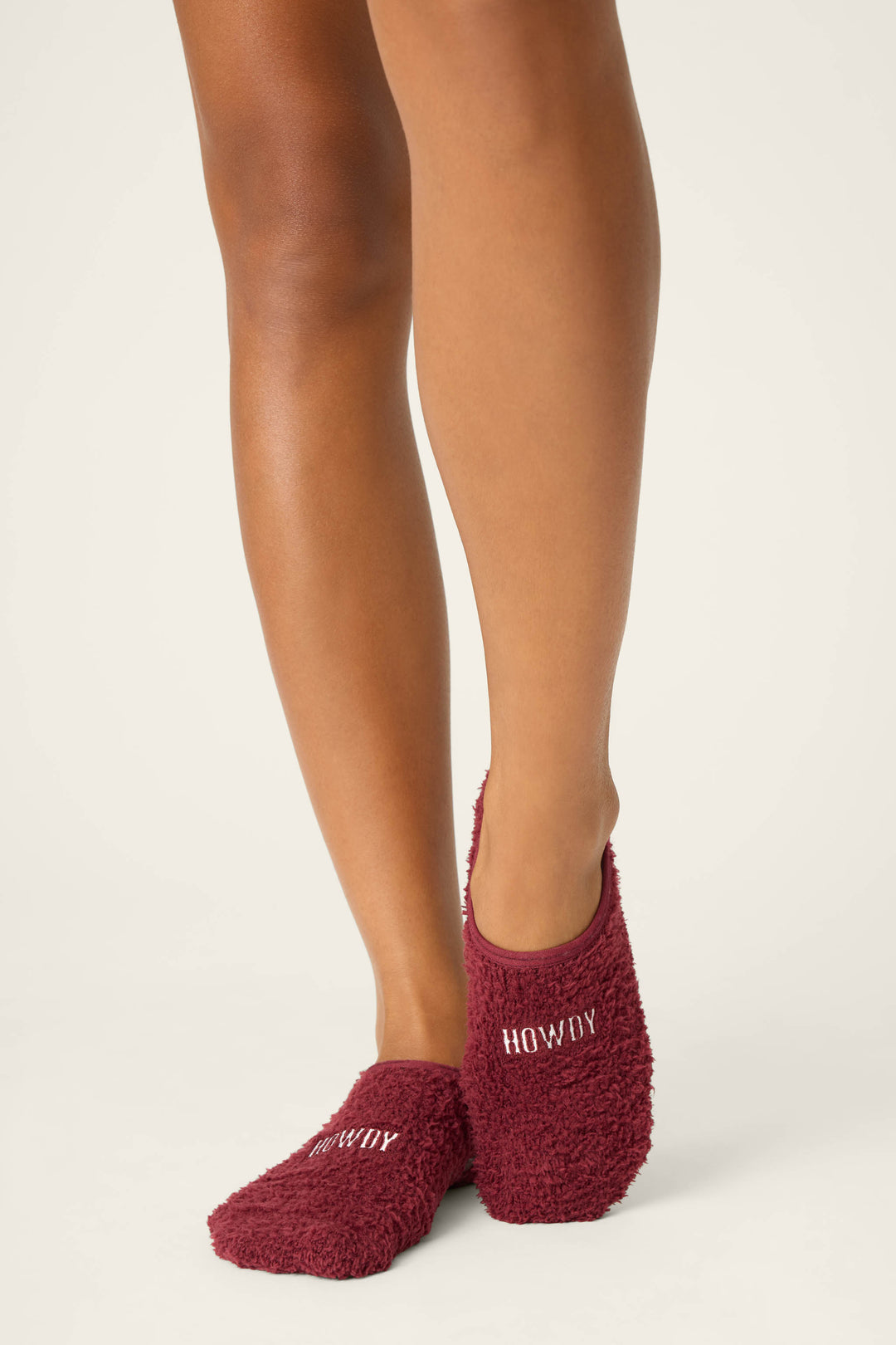 A person stands on tiptoe wearing P.J. Salvage check color - HOWDY SOCKS FLANNELS, featuring burgundy fuzzy slipper socks with HOWDY embroidered in white on one foot, set against a plain light background.