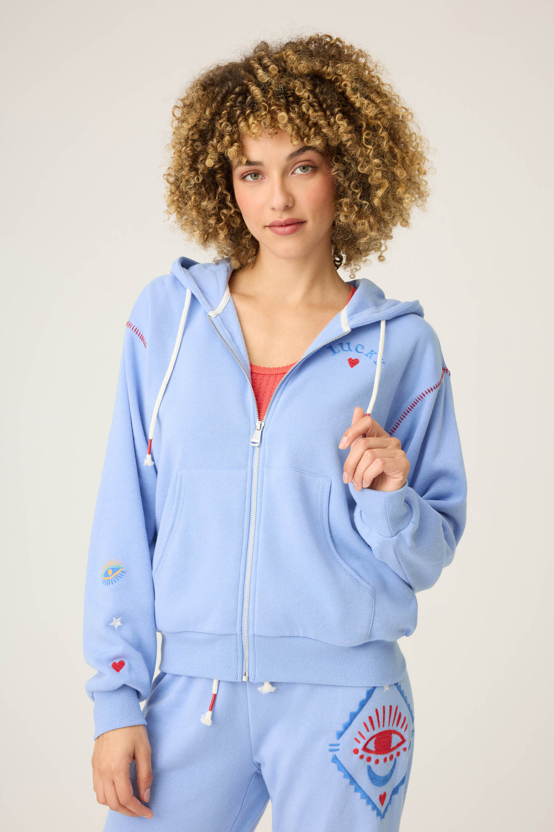 A woman with curly hair wears P.J. Salvage’s LUCKY HOODIE LADY LUCK, a light blue zip-up with white drawstrings and matching sweatpants featuring red and blue graphics, standing against a plain background holding the hoodie.