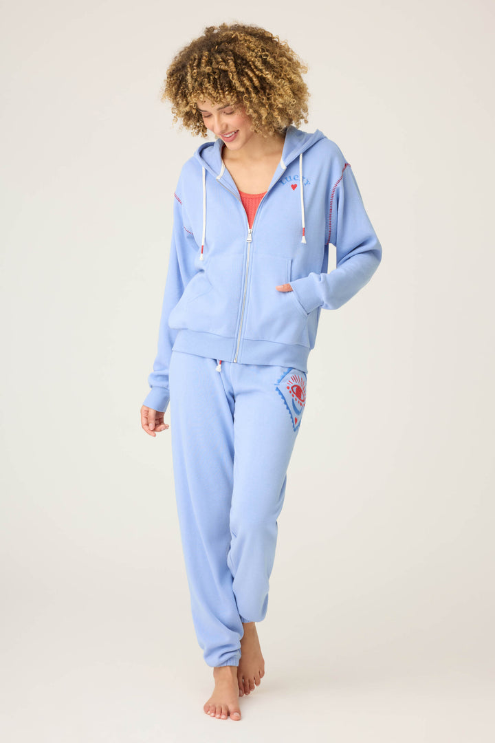 A person with curly hair wears the P.J. Salvage LUCKY HOODIE LADY LUCK in light blue, paired with matching sweatpants and a red top underneath. Barefoot, they stand on a plain background, looking down with a slight smile.