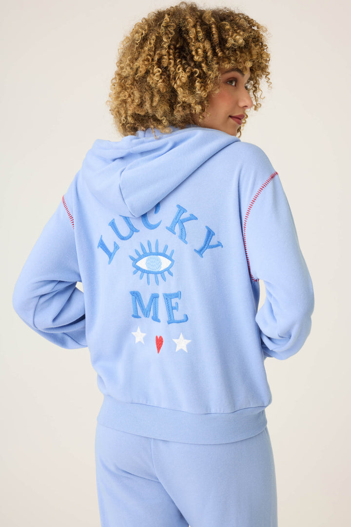 A person with curly hair models the P.J. Salvage LUCKY HOODIE LADY LUCK in light blue, featuring “LUCKY ME,” an eye graphic, two white stars, and a red heart on the back. They are turned slightly to the side, facing away from the camera.