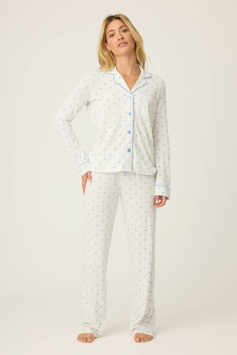 A woman stands barefoot in the P.J. Salvage Pant Set FLOCKING NOTCH COLLAR PJ SET LADY LUCK—a light pajama set with blue polka dots, blue trim, a long-sleeve button-up top, and full-length pants—against a plain light background.