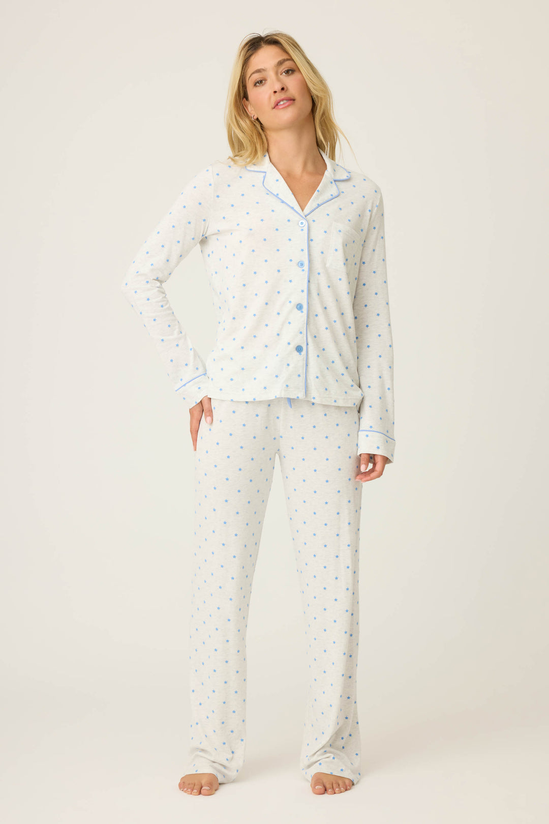 A woman stands barefoot in the P.J. Salvage Pant Set FLOCKING NOTCH COLLAR PJ SET LADY LUCK—a light pajama set with blue polka dots, blue trim, a long-sleeve button-up top, and full-length pants—against a plain light background.