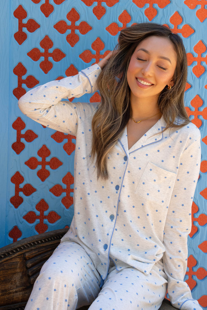 A woman wearing the P.J. Salvage Pant Set FLOCKING NOTCH COLLAR PJ SET LADY LUCK sits on a bench, smiling with her eyes closed and touching her hair, in front of a blue wall with an orange geometric pattern.