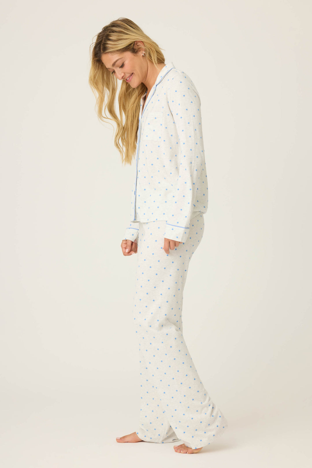 A woman with long, wavy blonde hair stands barefoot and slightly angled on a white background, wearing the P.J. Salvage Pant Set FLOCKING NOTCH COLLAR PJ SET LADY LUCK—light-colored pajamas with a blue dot pattern.
