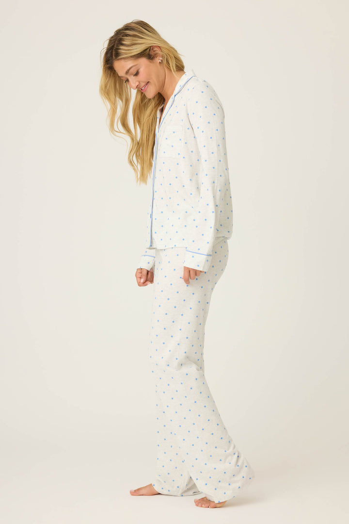 A woman with long, wavy blonde hair stands barefoot and slightly angled on a white background, wearing the P.J. Salvage Pant Set FLOCKING NOTCH COLLAR PJ SET LADY LUCK—light-colored pajamas with a blue dot pattern.