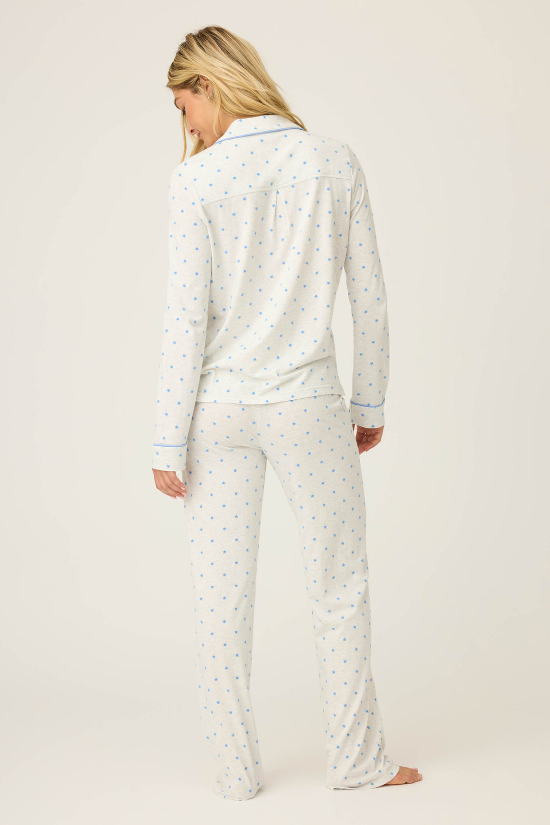 A person with long blonde hair stands barefoot facing away, wearing the P.J. Salvage Pant Set FLOCKING NOTCH COLLAR PJ SET LADY LUCK, featuring white pajamas with a small blue dot pattern, against a plain light background.