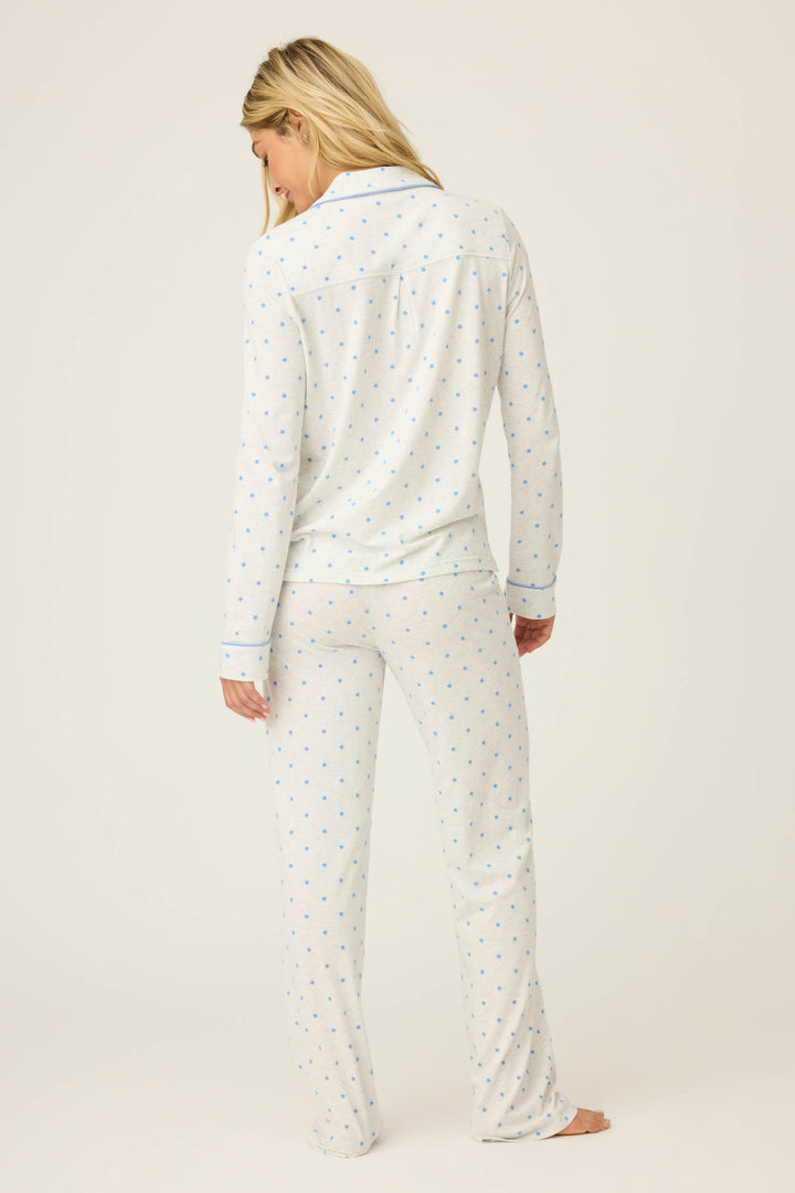A person with long blonde hair stands barefoot facing away, wearing the P.J. Salvage Pant Set FLOCKING NOTCH COLLAR PJ SET LADY LUCK, featuring white pajamas with a small blue dot pattern, against a plain light background.