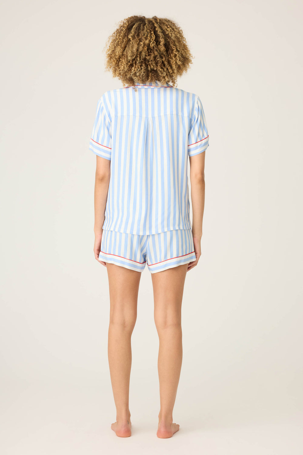 A person stands barefoot facing away from the camera, wearing the P.J. Salvage Short Set STRIPE PJ SET LADY LUCK— a matching short-sleeve shirt and shorts with vertical blue and white stripes and red trim accents.