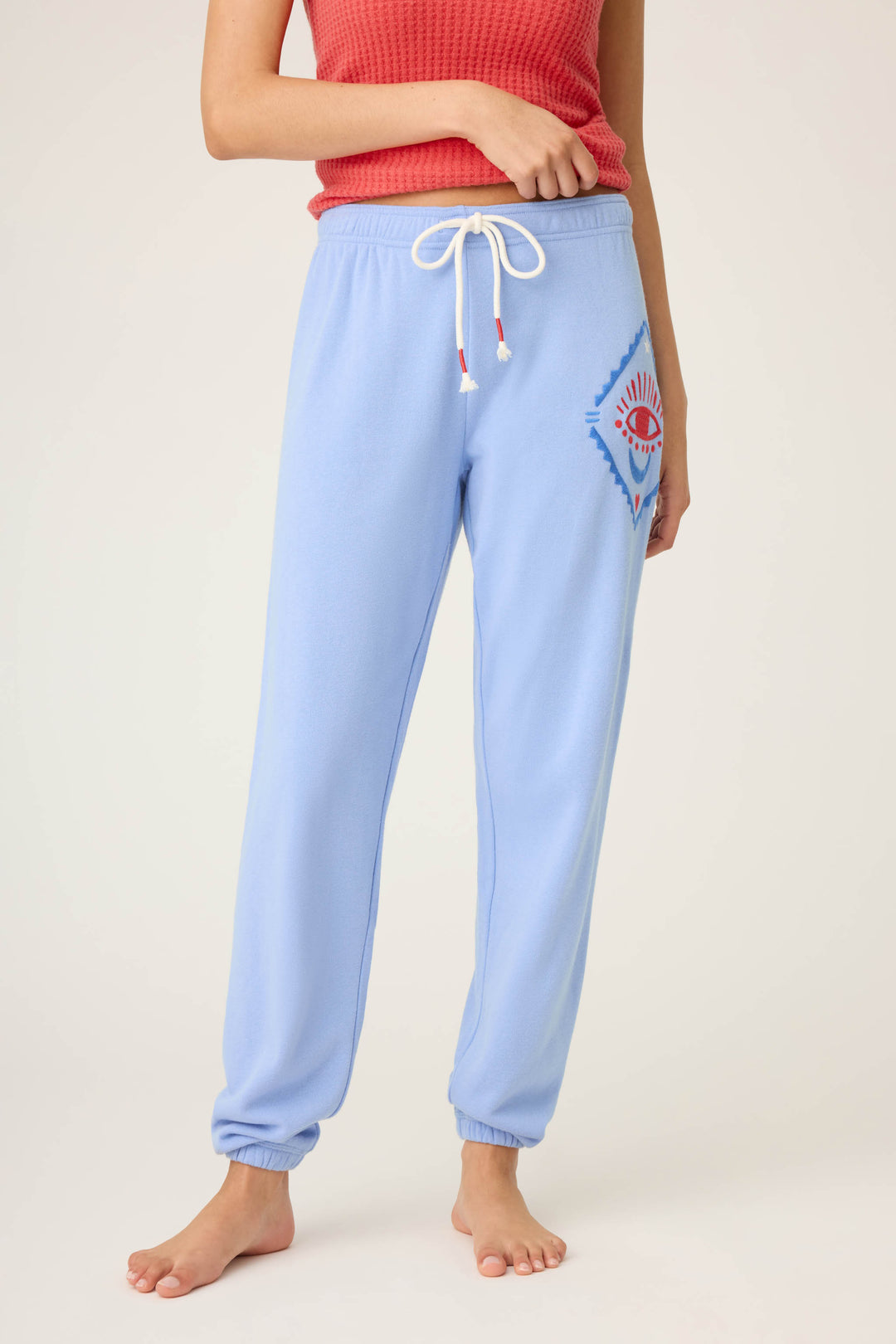 A person wearing the P.J. Salvage SOLID BANDED PANT LADY LUCK—light blue jogger sweatpants with a white drawstring and a colorful embroidered eye graphic—standing barefoot against a plain background in a sleeveless red textured top.