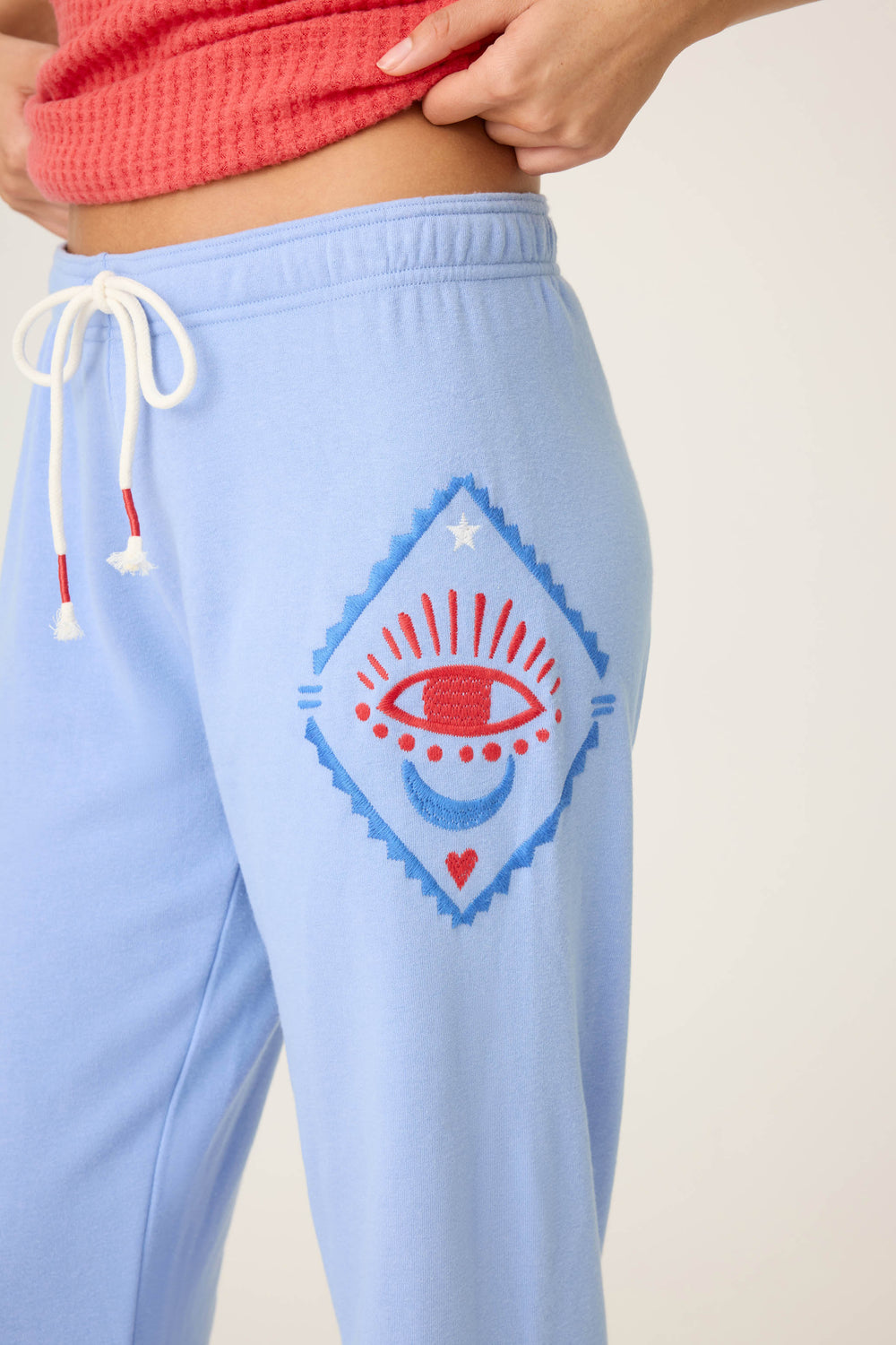 A person wears P.J. Salvage SOLID BANDED PANT LADY LUCK in light blue, featuring a white drawstring and red/blue eye design with geometric shapes, star, and heart on the upper thigh, paired with a red top.
