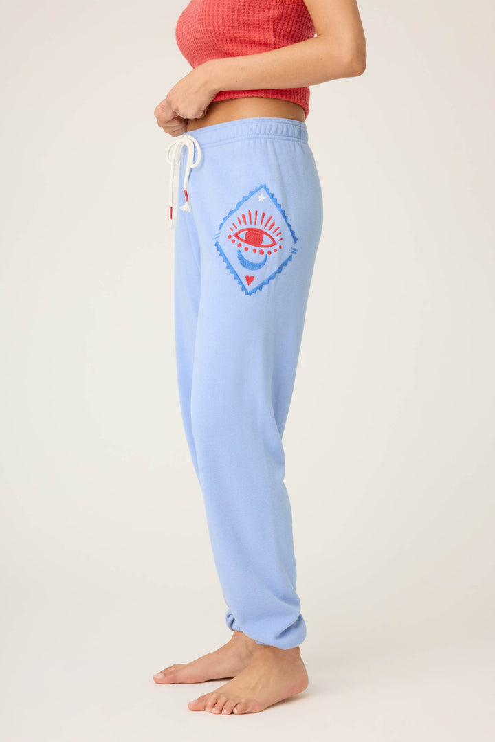 A person stands sideways barefoot, wearing P.J. Salvages SOLID BANDED PANT LADY LUCK—light blue sweatpants featuring a red and blue eye design inside a diamond on the side—with a red textured top.