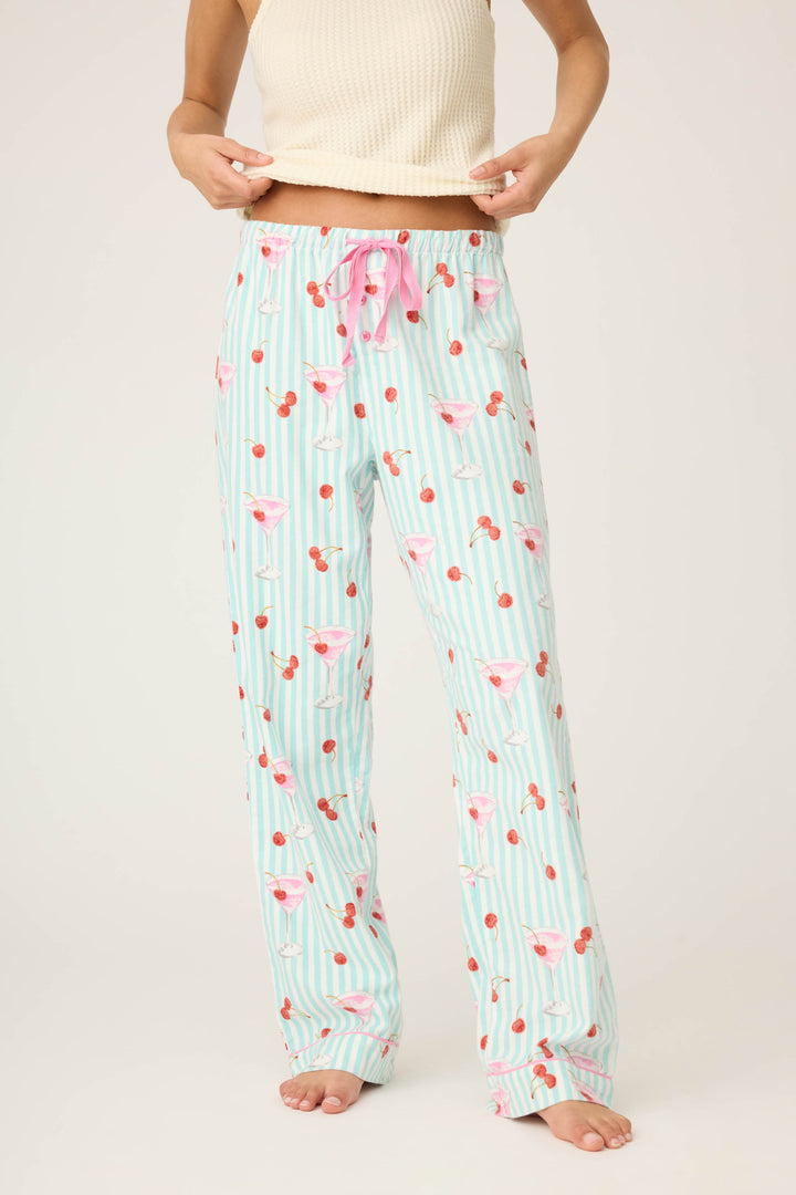 A person wears P.J. Salvage’s DOGS PANT FLANNELS—light blue and white striped pajama pants with pink flamingos and cherries—paired with a cream sleeveless top, barefoot, cropped from shoulders down.