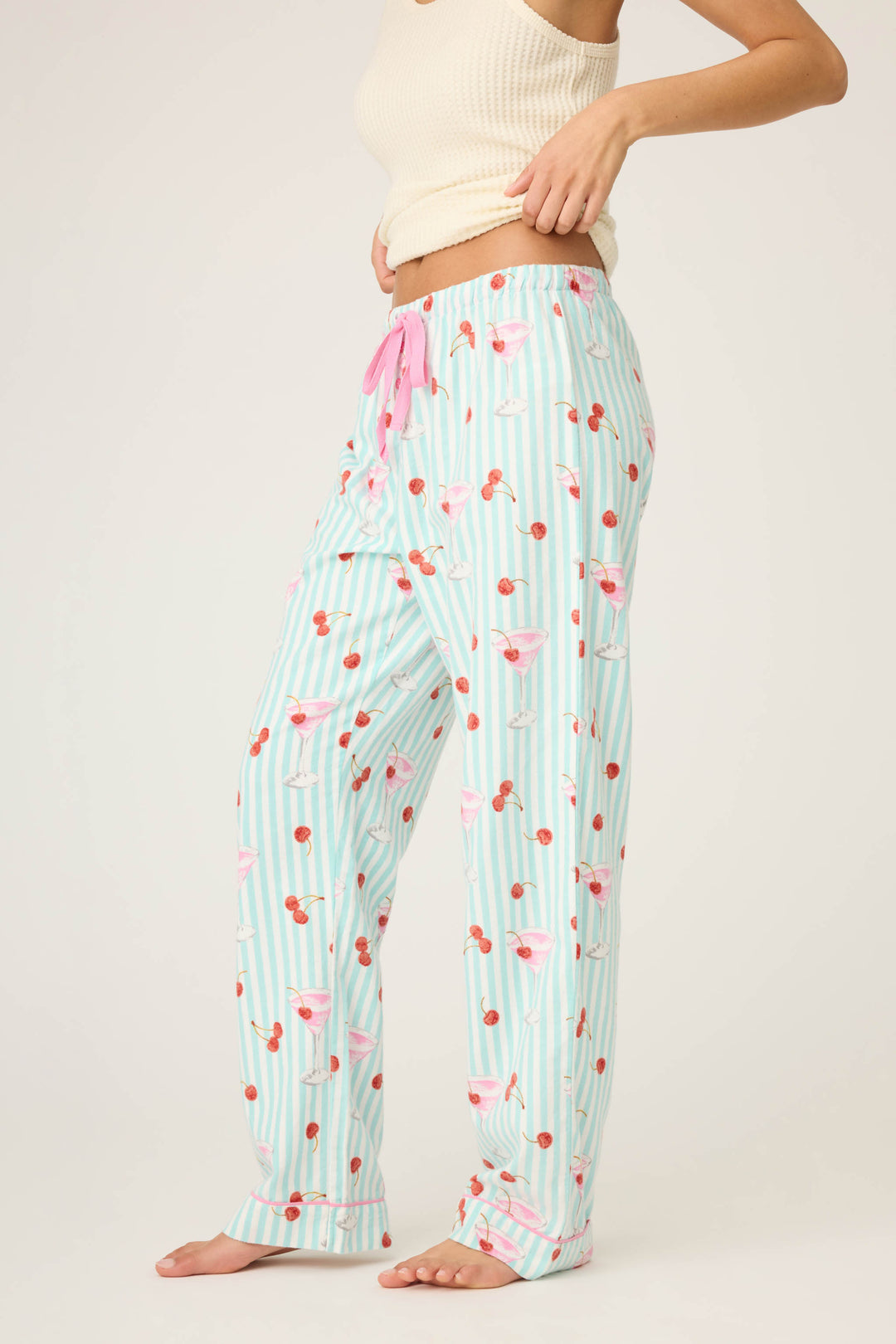 A person wears P.J. Salvage DOGS PANT FLANNELS, light blue pajama pants with a pink drawstring and fun prints, plus a cream ribbed tank top. Theyre barefoot, standing sideways against a plain background.