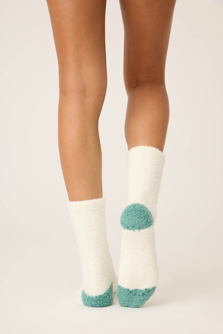 A person stands on tiptoes, showing the back of their lower legs and feet in P.J. Salvage Cheers Fuzzy Crew Socks—white with teal accents at the heels and toes—against a plain light background.