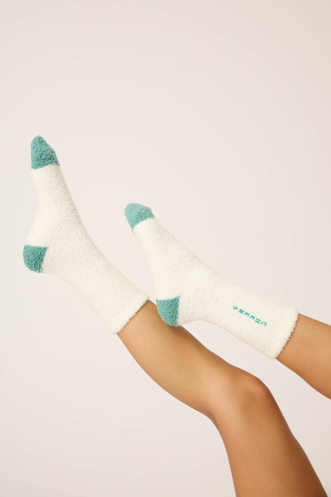 A pair of legs wears P.J. Salvage Cheers Fuzzy Crew Socks—white with teal accents on toes, heels, and cuffs—against a plain light background. "Cheers" is printed on the side.
