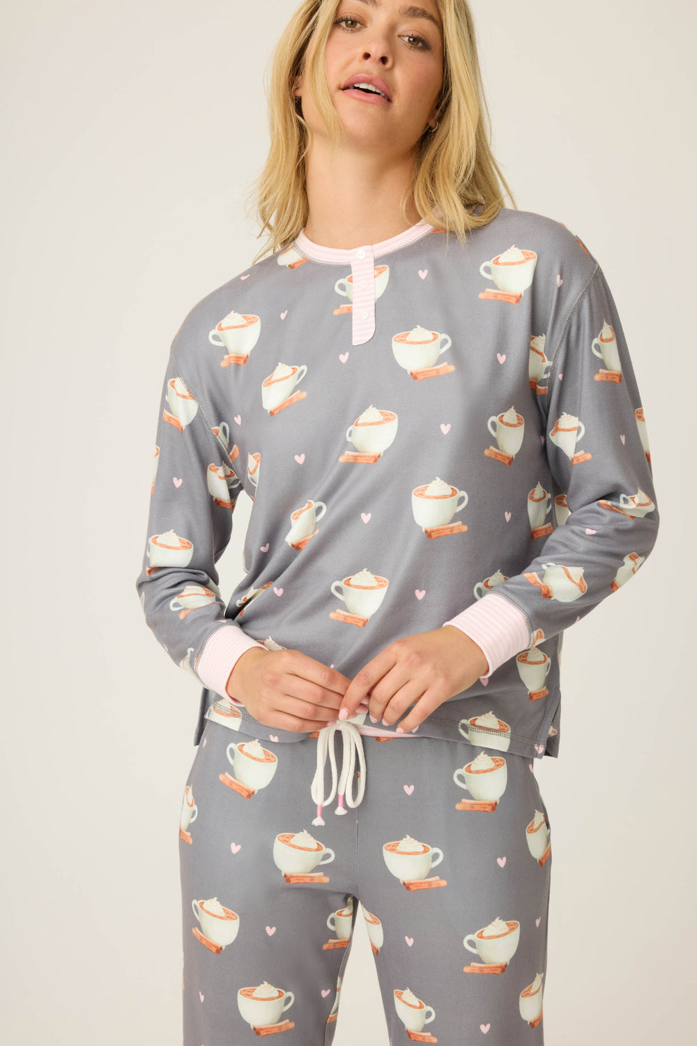 A woman wears the P.J. Salvage Love You A Latte Coffee Print long sleeve top with matching gray pajama pants, featuring cup and heart prints, pink accents at the cuffs and neckline, and pants with a white drawstring.