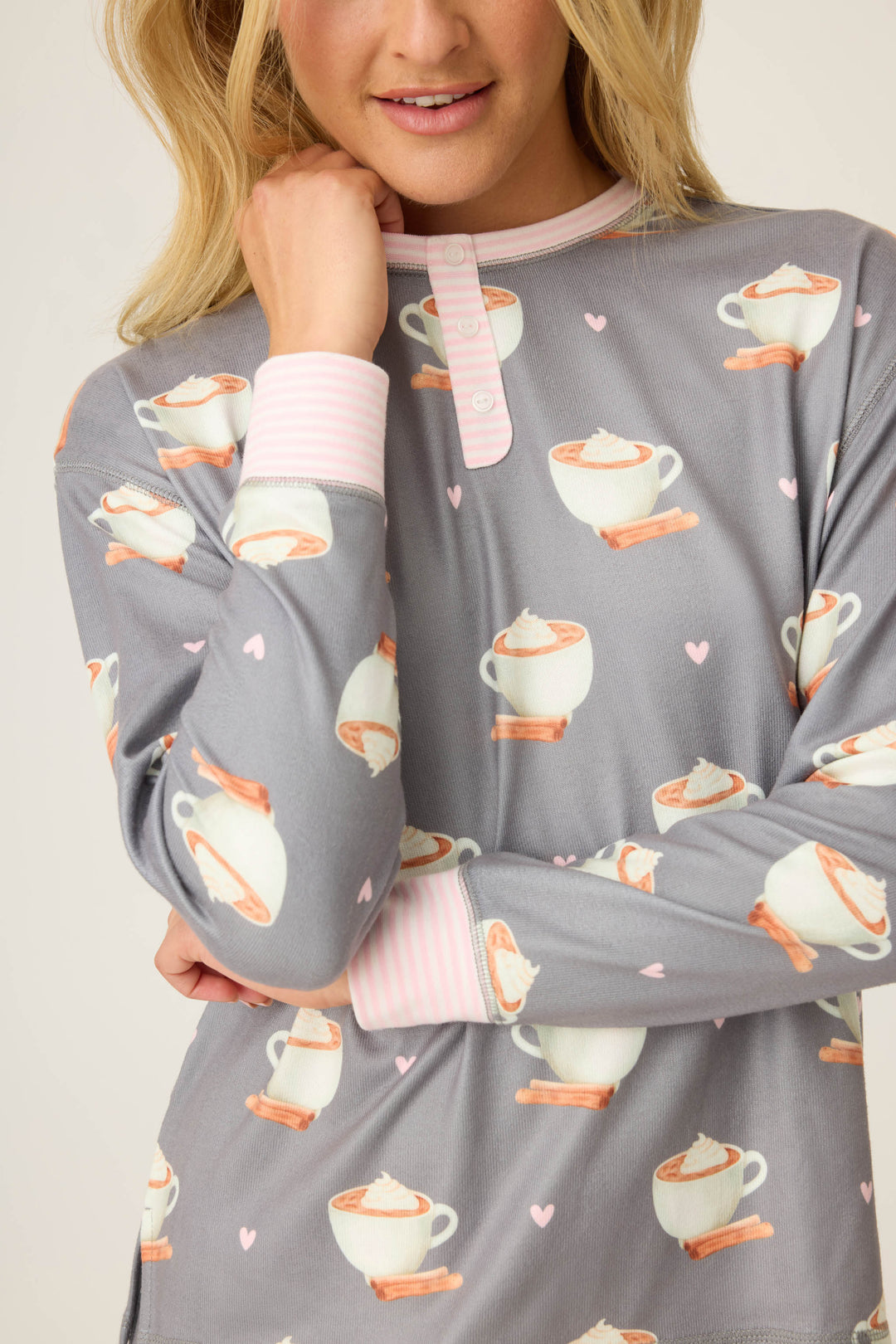 A woman wears the P.J. Salvage Love You A Latte Coffee Print long sleeve top, featuring mugs with whipped cream, cinnamon sticks, pink hearts, and pink-and-white striped collar and cuffs. Her hand rests near her chin.