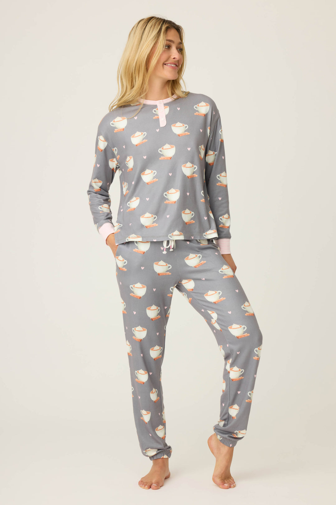 A person wears the P.J. Salvage Love You A Latte Coffee Print Long Sleeve Top and matching pants, featuring a gray base with white teacups. They stand with one hand in a pocket, smiling and looking slightly to the side.