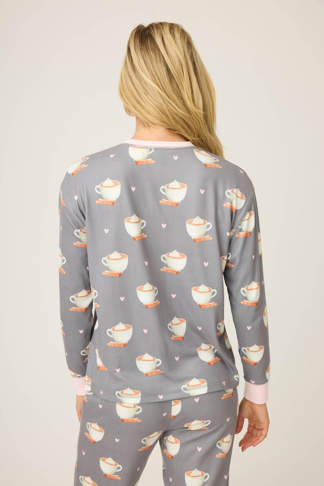 A person with long blonde hair is shown from the back wearing the P.J. Salvage Love You A Latte Coffee Print Long Sleeve Top, featuring a teacup and pink heart pattern. The background is plain and light-colored.