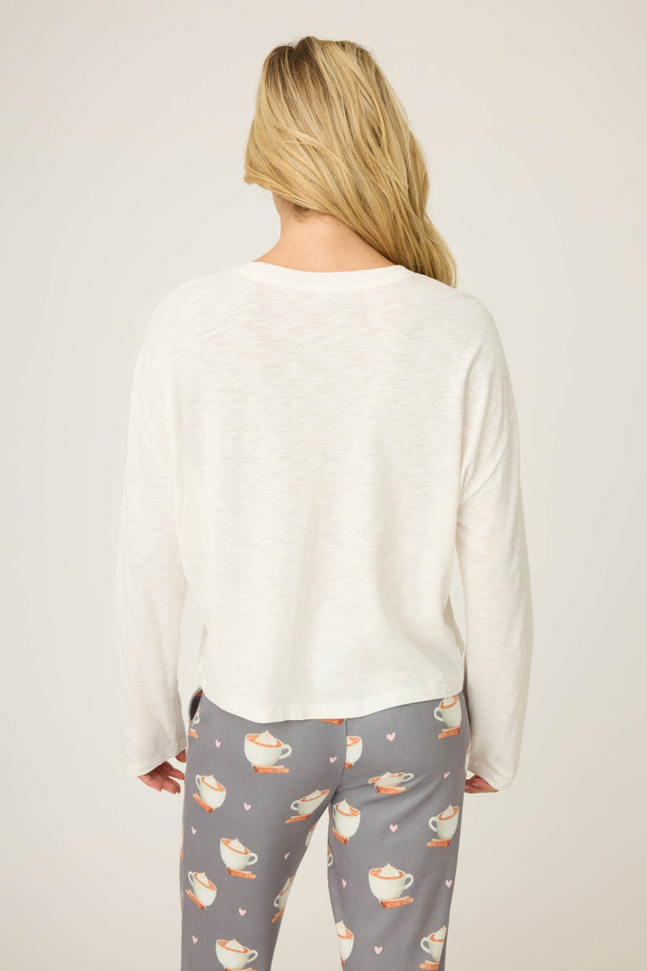 A person with long blonde hair is seen from the back wearing the P.J. Salvage Love You A Latte Coffee Print Long Sleeve Top and gray pajama pants with teacup and heart patterns, against a plain, light background.