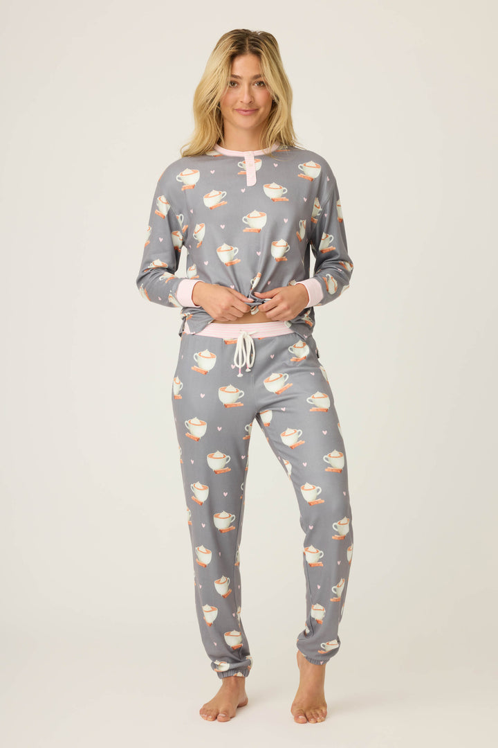 A person stands barefoot against a plain background, wearing P.J. Salvages Love You A Latte Coffee Print Banded Pant pajama set with matching gray fabric, cup prints, pink collar and cuff accents, and a drawstring waist.