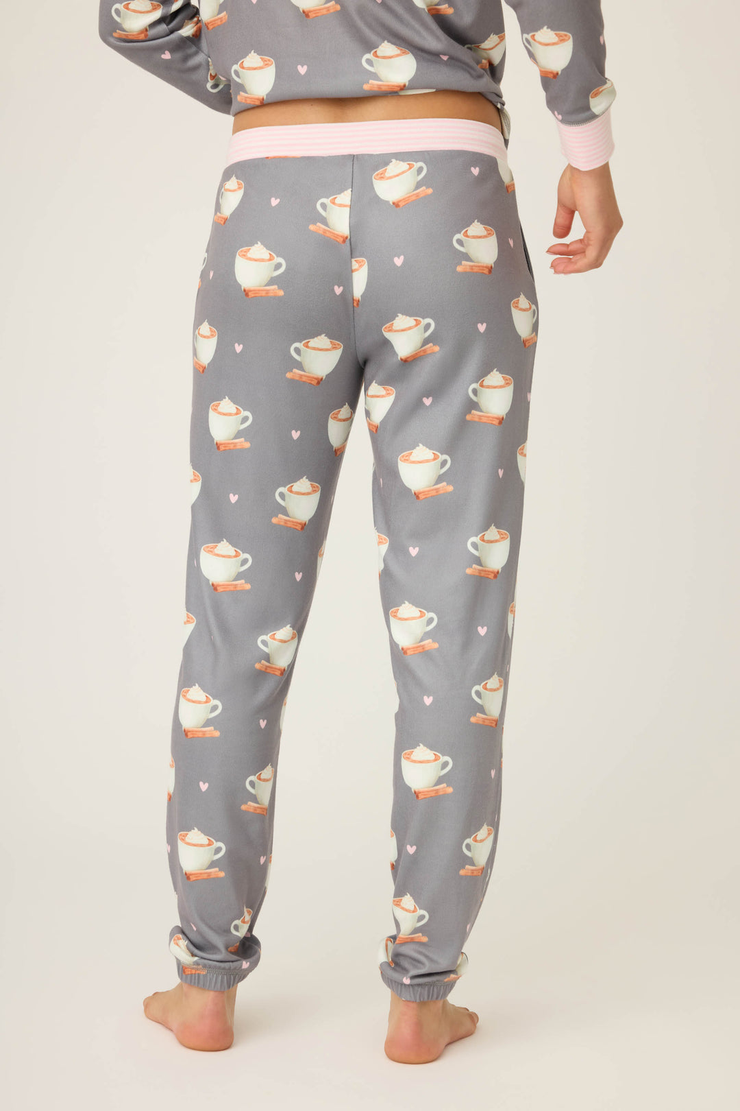 A person stands barefoot, shown waist down, wearing P.J. Salvages Love You A Latte Coffee Print Banded Pant—gray pajama pants with a cup and saucer pattern and pink hearts. Their hand is relaxed at their side.