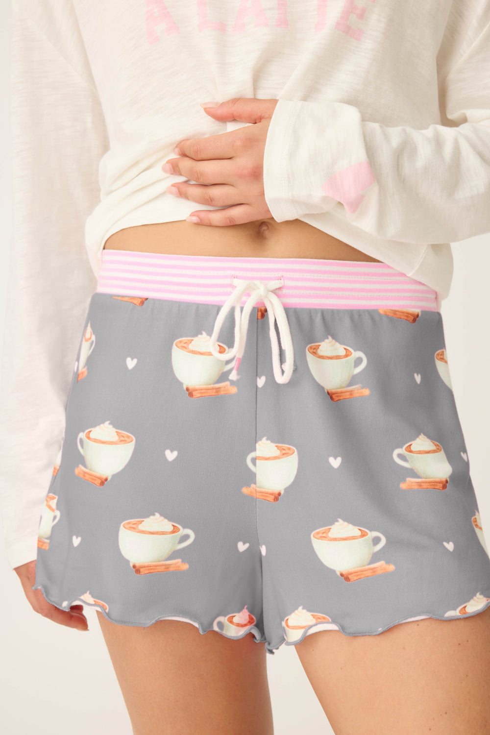 A person wears P.J. Salvage Love You A Latte Coffee Print Shorts featuring mugs of hot chocolate, cinnamon sticks, and hearts, with a pink and white striped waistband and drawstring, paired with a white long-sleeve top.